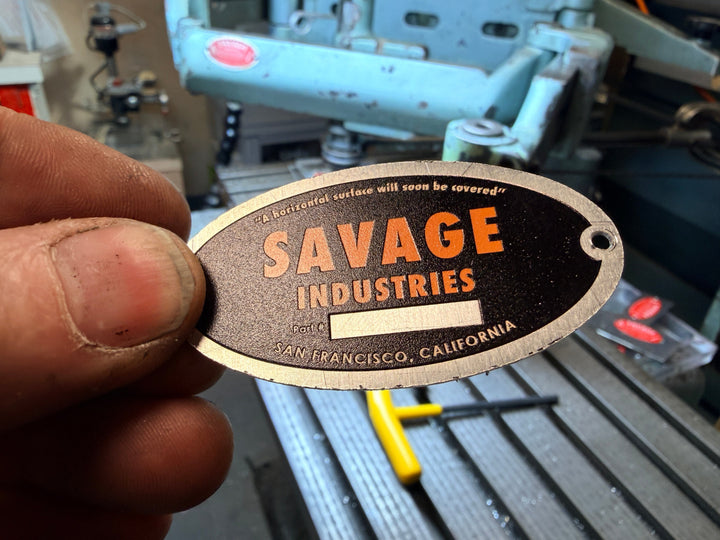 Hand holding a Savage Industries metal tag in an industrial setting