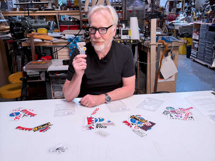 Adam Savage holding a mini spacesuit sticker in a workshop with other Mega Sticker Packs on the table #hero #desktop